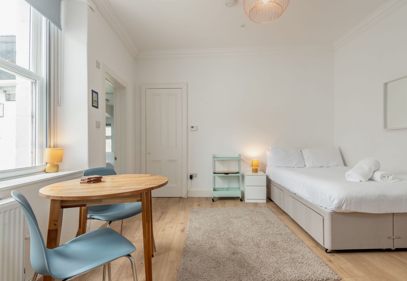 Studio with dining table, blue chairs, double bed, and large window letting in natural light.