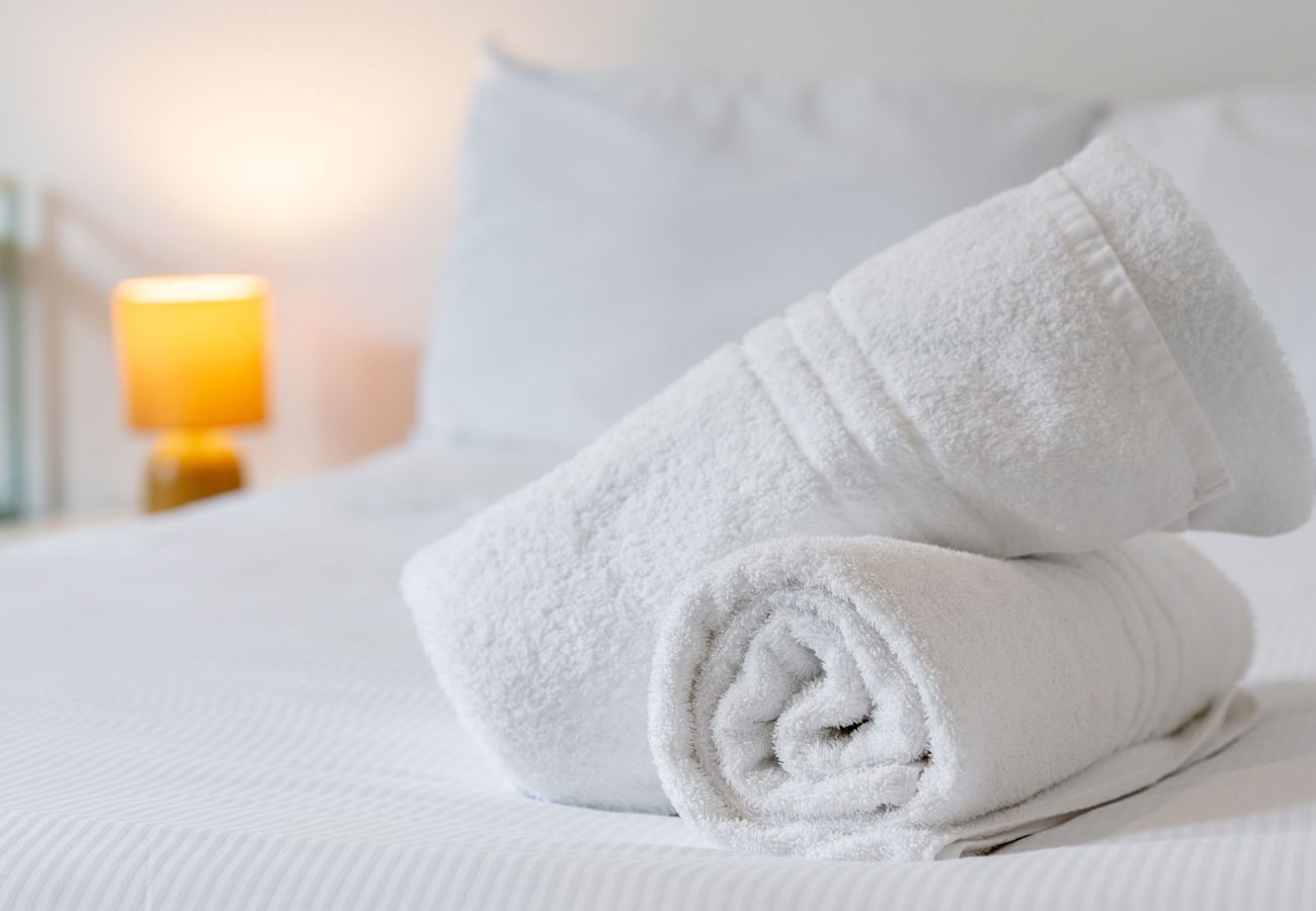 Two rolled white towels on a bed next to a lit bedside lamp.