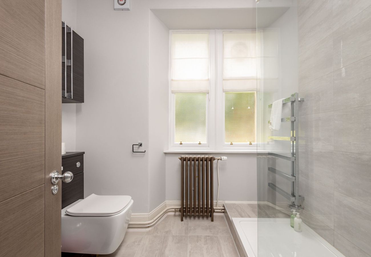 Contemporary bathroom featuring walk-in glass shower, wall-mounted toilet, and twin windows with privacy blinds.