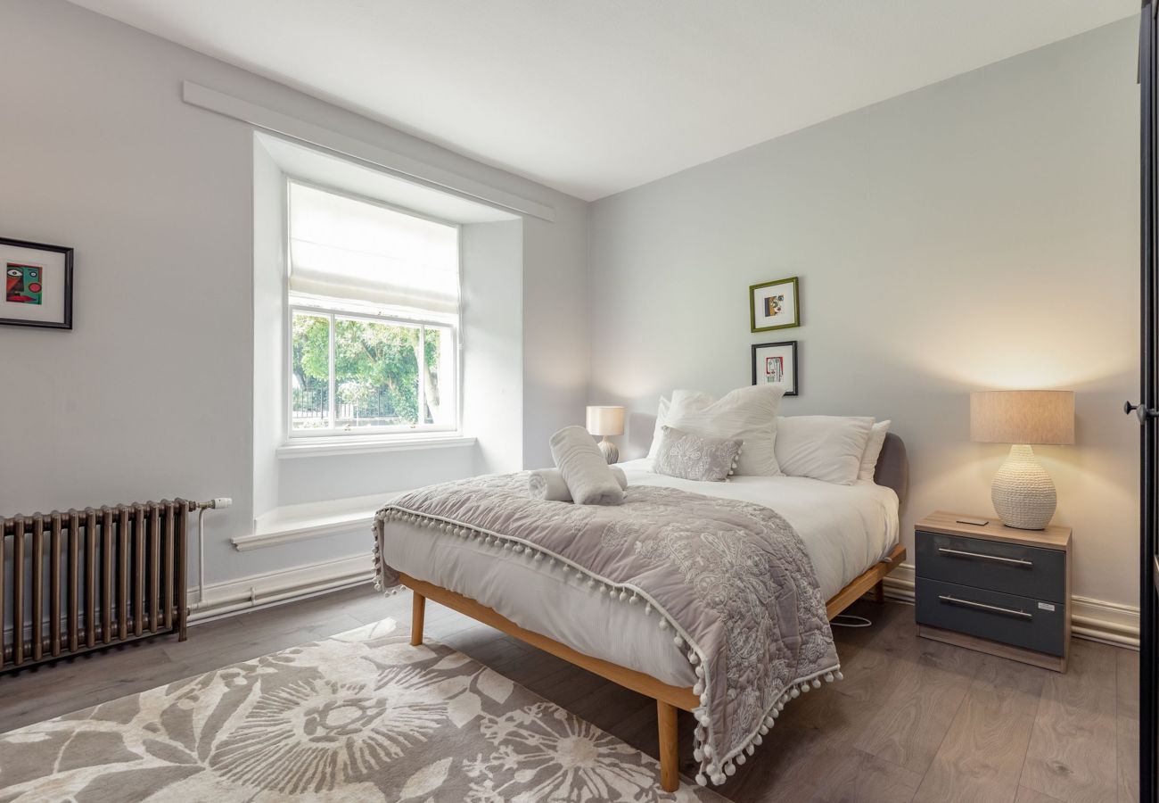 Peaceful double bedroom with soft grey walls, neatly made bed with pom-pom throw, bedside lamps, and a view to the garden.
