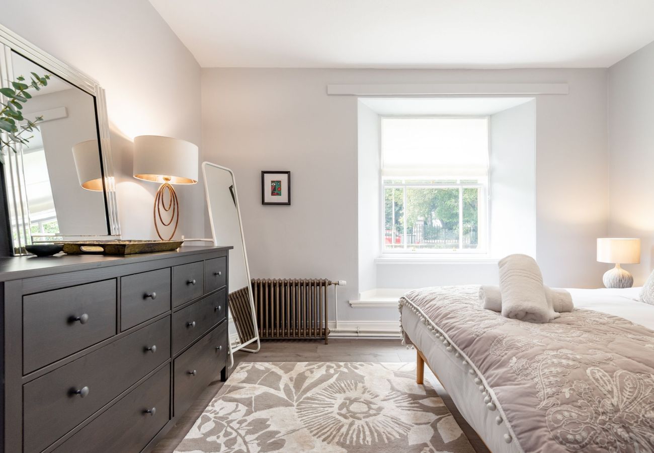 Serene double bedroom with soft lighting, floor mirror, large dresser, and a bright window looking out to trees.