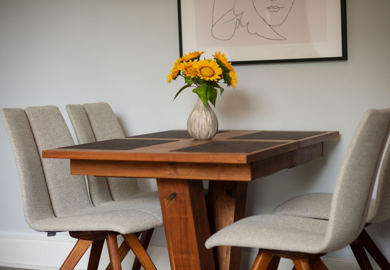 Square wooden dining table with grey upholstered chairs, topped with a vase of sunflowers and framed artwork above.