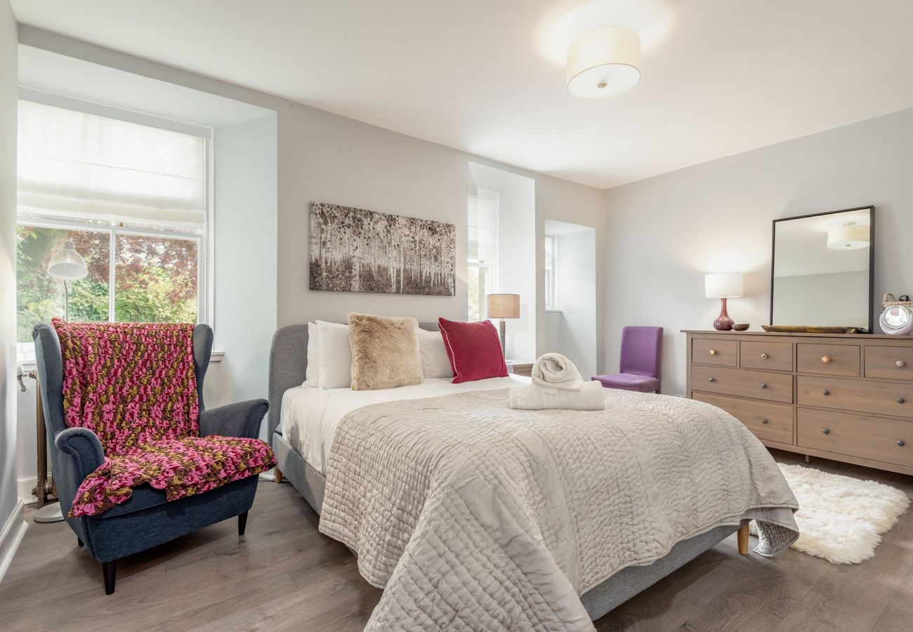 Spacious double bedroom featuring a grey upholstered bed, large dresser, bright accent pillows, and twin windows with leafy views.