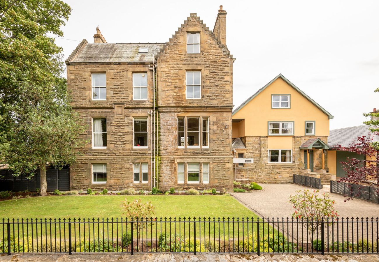 Straight-on view of sandstone apartment building with mature trees, gated entrance, and neat formal lawn.