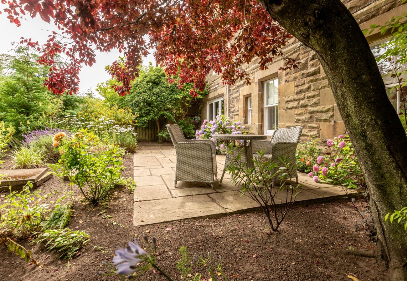  Tranquil garden patio with wicker table and chairs beneath a deep red maple tree, surrounded by flowering plants.
